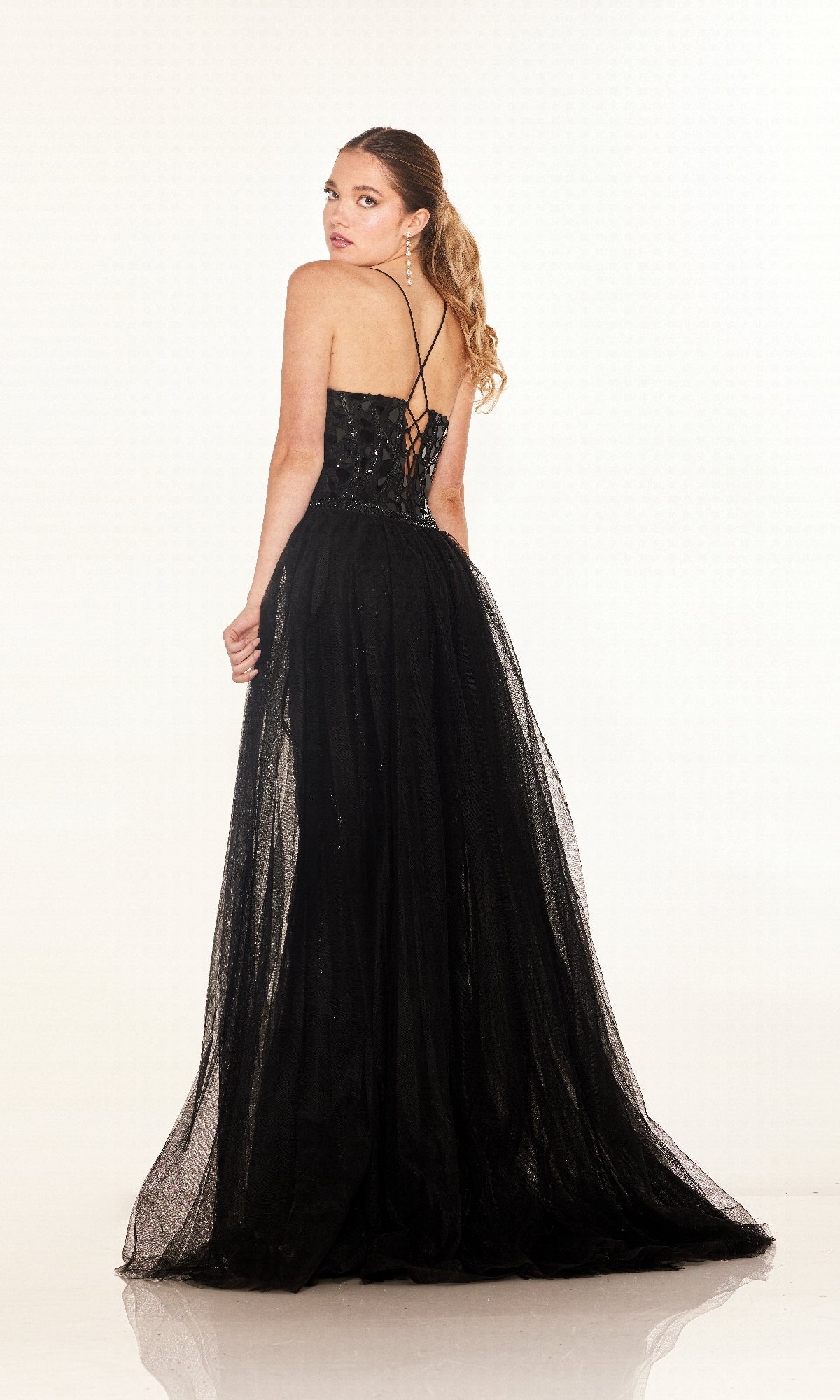 A woman with long hair in a ponytail models the Cecilia Couture C132, a floor-length black prom dress with thin straps, fitted bodice, and lace-up back, standing slightly turned to showcase the design.